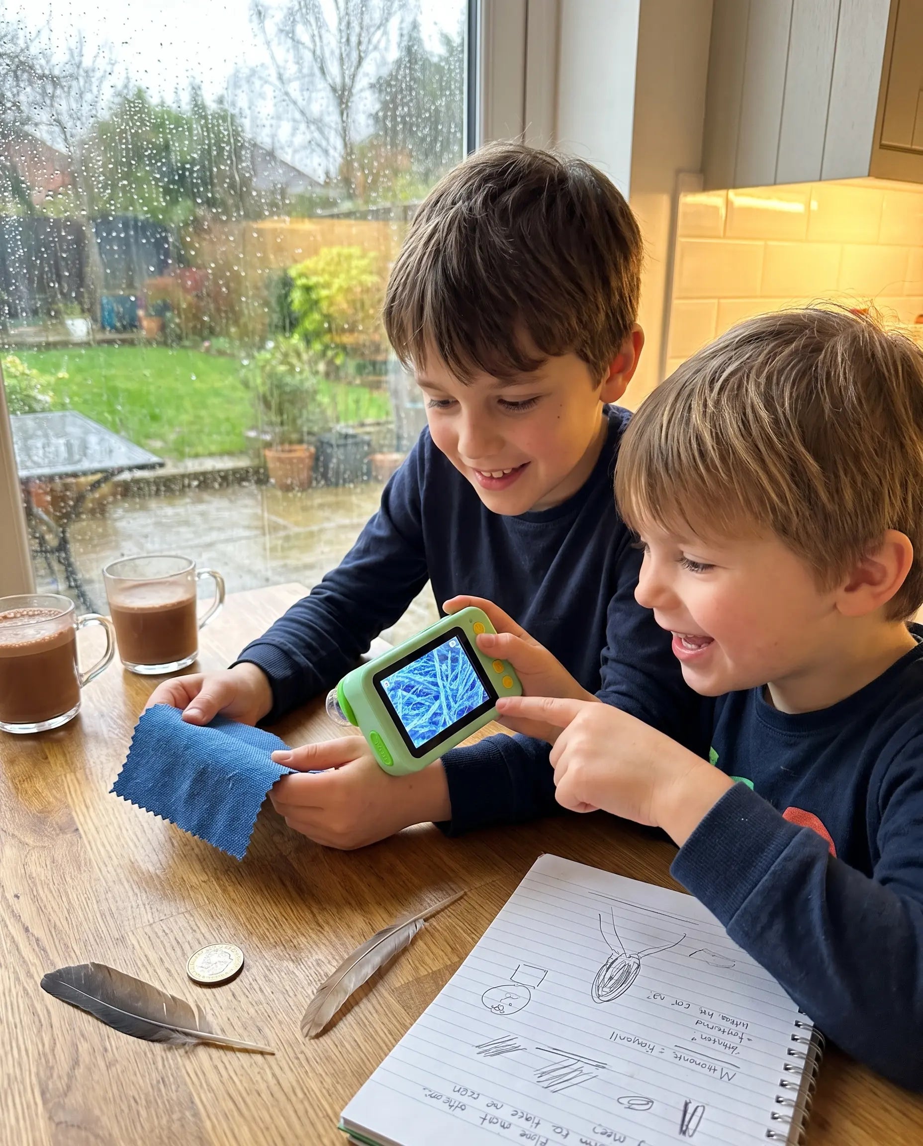 Two smiling siblings using the Discovery Microscope at a kitchen table on a rainy day
